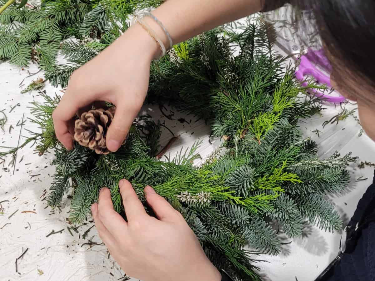 A woman crafts a wreath using pine cones, showcasing her creativity and attention to detail in the arrangement.