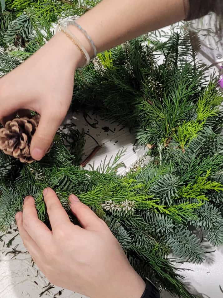 A woman crafts a wreath using pine cones, showcasing her creativity and attention to detail in the arrangement.
