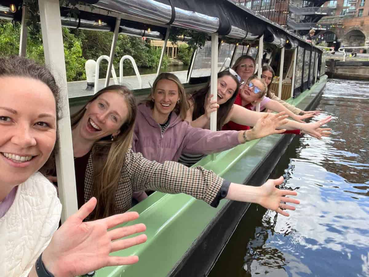 A team of women on a boat at a London canal lock, actively participating in a game, with their hands raised in unison.