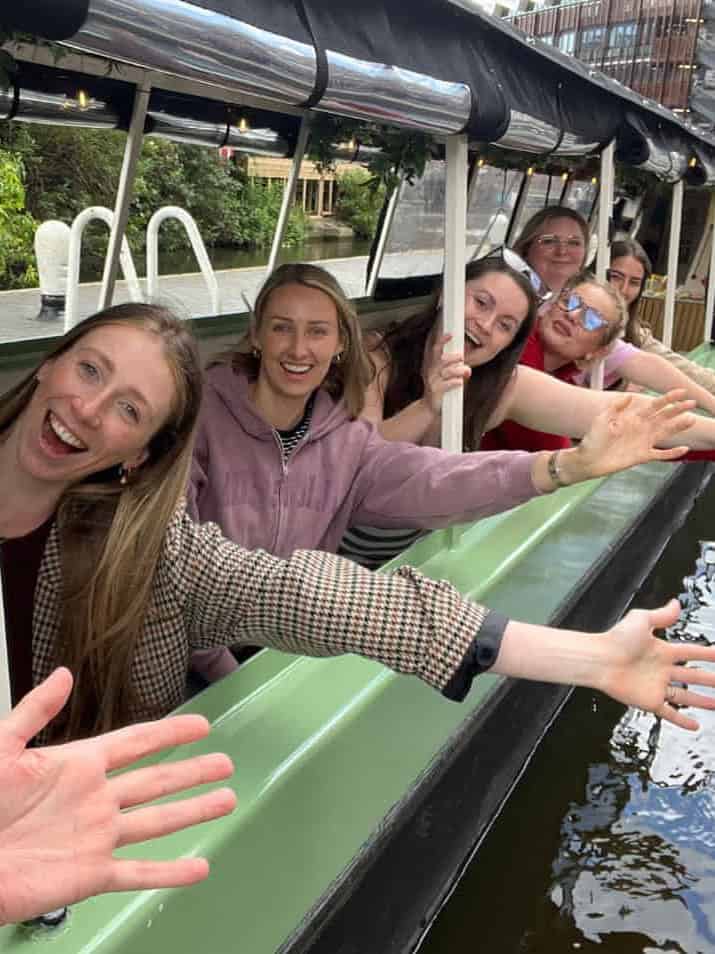 A team of women on a boat at a London canal lock, actively participating in a game, with their hands raised in unison.